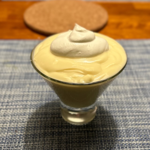 A short V shaped class is seen filled with a light yellow/tan colored pudding, and topped with a small dollop of whipped cream.